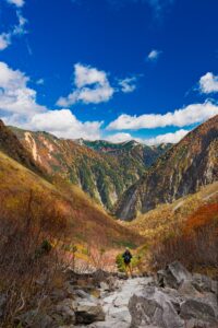 A hiker on a rocky trail in an autumn alpine valley beneath jagged peaks in the Japanese Alps, fine art landscape by Mitsuki