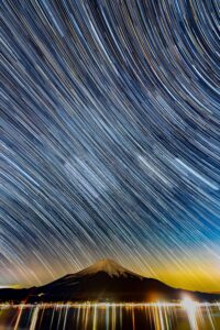 Star trails streaking above a snow-capped Mount Fuji with city lights below in central Japan, fine art photograph by Mitsuki